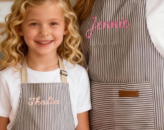 Puede incluir: Dos delantales, uno para adultos y otro para niños, con un estampado de rayas blancas y negras. El delantal para adultos tiene el nombre "Jennie" bordado en rosa, y el delantal para niños tiene el nombre "Thalia" bordado en rosa. Ambos delantales tienen una correa para el cuello beige y un bolsillo delantero.