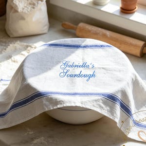 Puede incluir: Un cuenco blanco cubierto con un paño blanco y azul a rayas, con el texto "Gabriella's Sourdough" en azul. Una bolsa de harina y un rodillo están en el fondo. La escena sugiere hornear.
