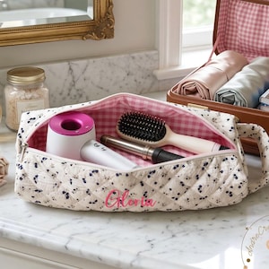 May include: A floral quilted cosmetic bag with the name "Gloria" embroidered in pink. The bag is open, revealing a hairdryer, brush, and other beauty tools. A vintage suitcase and toiletries are in the background.