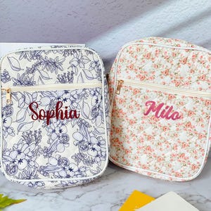 Può includere: Due borse a tracolla trapuntate. Una borsa presenta uno sfondo bianco con un motivo floreale blu e il nome "Sophia" ricamato in rosso. L'altra borsa ha uno sfondo bianco con un motivo floreale rosa e il nome "Milo" ricamato in rosa. Entrambe le borse hanno cerniere dorate.
