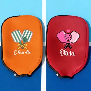 Puede incluir: Dos fundas para palas de pickleball, una naranja y otra roja, se muestran sobre un fondo azul. La funda naranja tiene un diseño de pala a rayas verdes y blancas con el nombre "Charlie". La funda roja presenta un diseño de pala rosa con el nombre "Olivia".