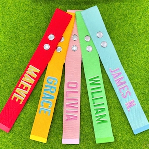 Op de afbeelding: Vijf kleurrijke stoffen banden met geborduurde namen in contrasterende kleuren. De banden zijn rood, geel, roze, groen en lichtblauw. Elke band heeft twee zilveren klinknagels. De geborduurde namen zijn Maeve, Grace, Olivia, William en James N.