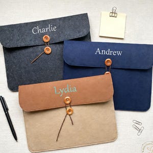 May include: Three personalized felt laptop sleeves in gray, navy blue, and tan. Each sleeve features a name embroidered in a contrasting color and fastens with a wooden button and loop closure. A black pen and sticky note are also visible.