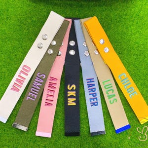 Op de afbeelding: Een verzameling gepersonaliseerde canvas riemen in verschillende kleuren, elk geborduurd met een naam of initialen. De riemen hebben zilverkleurige metalen hardware en worden getoond op een groene ondergrond. Kleuren zijn crème, olijf, roze, marineblauw, lichtblauw, beige en geel.