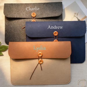 May include: Three personalized felt laptop sleeves in gray, navy blue, and tan. Each sleeve features a name embroidered on the flap: "Charlie", "Andrew", and "Lydia". The sleeves have a button closure and a brown string detail.