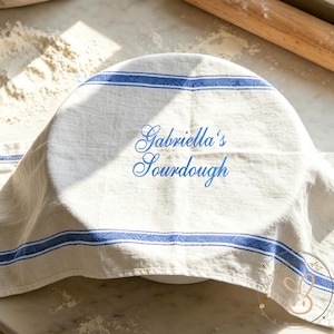 May include: A white bowl covered with a beige linen cloth with blue stripes and the text "Gabriella's Sourdough" in blue script. The cloth is draped over the bowl, suggesting a baking or food preparation theme.