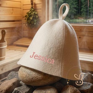 Könnte beinhalten: Eine cremefarbene Filz-Saunamütze mit einer Aufhängeschlaufe an der Oberseite, auf deren Vorderseite der Name Jessica in rosa Garn gestickt ist. Die Mütze liegt auf einem Haufen dunkelgrauer Steine in einer Holzsauna mit einem Holzeimer und einer Kelle.