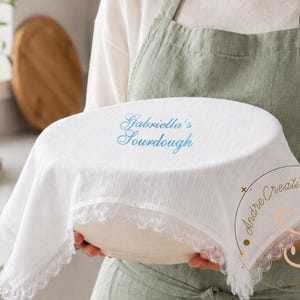 Puede incluir: Una funda de tela blanca para cuenco con un borde de encaje delicado, que presenta el texto bordado en azul Gabriella's Sourdough en la parte superior. La funda est&aacute; colocada sobre un cuenco redondo, mostrando un material texturizado y ligero.