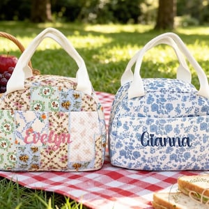 Pode incluir: Duas lancheiras personalizadas com alças brancas sobre uma toalha de piquenique xadrez vermelha e branca. Uma bolsa é patchwork com o nome "Evelyn" bordado em rosa. A outra é floral azul com "Gianna" em azul.