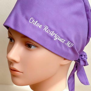 May include: A lilac surgical scrub cap with the embroidered name "Chloe Rodriguez MD". The cap is tied at the back with matching fabric ties. The cap is displayed on a mannequin head.