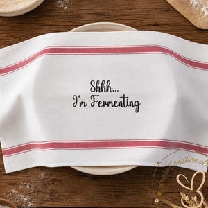 Puede incluir: Paño de cocina blanco con rayas rojas y el texto "Shhh... I'm Fermenting". El paño está colocado sobre un plato blanco sobre una superficie de madera. También se ven harina y utensilios de cocina.