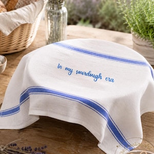 May include: A white linen bread cloth with blue stripes and the embroidered text "in my sourdough era" covers a bowl. A basket of bread, lavender, and flour are also visible, suggesting a baking theme.