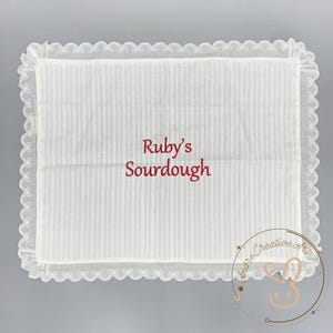 Puede incluir: Un pa&ntilde;o rectangular de tela blanca con un sutil patr&oacute;n de rayas verticales y un borde decorativo de encaje festoneado. El texto Ruby's Sourdough est&aacute; bordado en el centro en una fuente cursiva de color rojo oscuro. La tela es semitransparente.