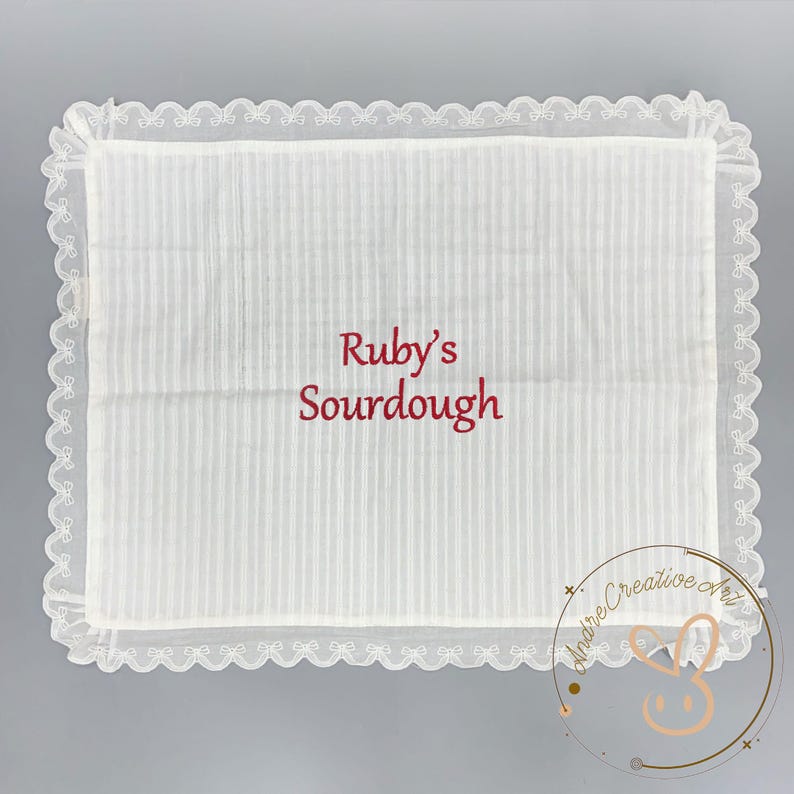 Puede incluir: Un pa&ntilde;o rectangular de tela blanca con un sutil patr&oacute;n de rayas verticales y un borde decorativo de encaje festoneado. El texto Ruby's Sourdough est&aacute; bordado en el centro en una fuente cursiva de color rojo oscuro. La tela es semitransparente.