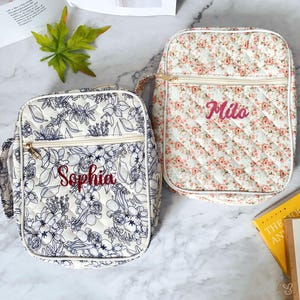 May include: Two quilted lunch bags. One bag features a white background with a blue floral design and the name "Sophia" embroidered in red. The other bag has a white background with a pink floral pattern and the name "Milo" embroidered in pink.