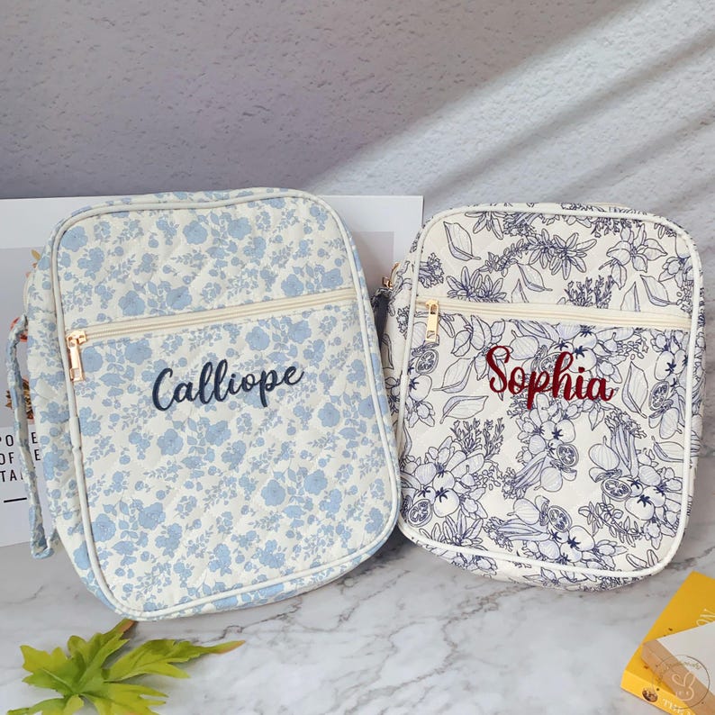 May include: Two floral-patterned bags with zippered closures. One bag features a light blue floral design with the name "Calliope" embroidered in navy. The other bag has a white background with a dark blue floral pattern and the name "Sophia" embroidered in red.