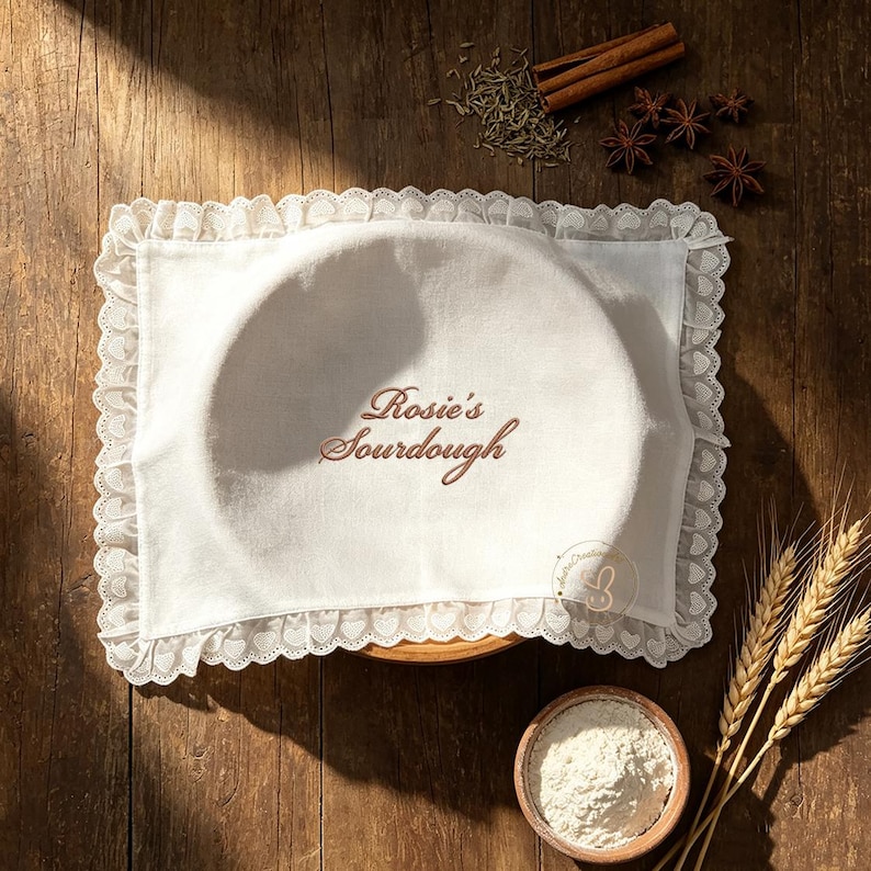 Puede incluir: Una funda de tela blanca para cestas de fermentaci&oacute;n con borde de encaje fruncido, que presenta el texto bordado en marr&oacute;n Rosie's Sourdough. La funda cubre un cuenco redondo, junto a un peque&ntilde;o recipiente con harina y especias.