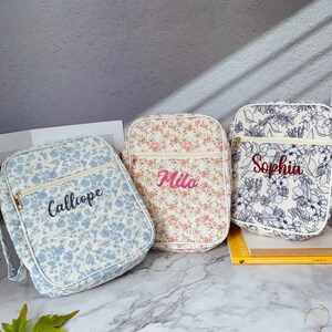 May include: Three floral pattern bags with personalized names. The bags feature a rectangular shape with a zippered front pocket and a top zipper. The bag on the left has a blue floral pattern with the name "Calliope". The middle bag has a pink floral pattern with the name "Mila". The bag on the right has a white and blue floral pattern with the name "Sophia".