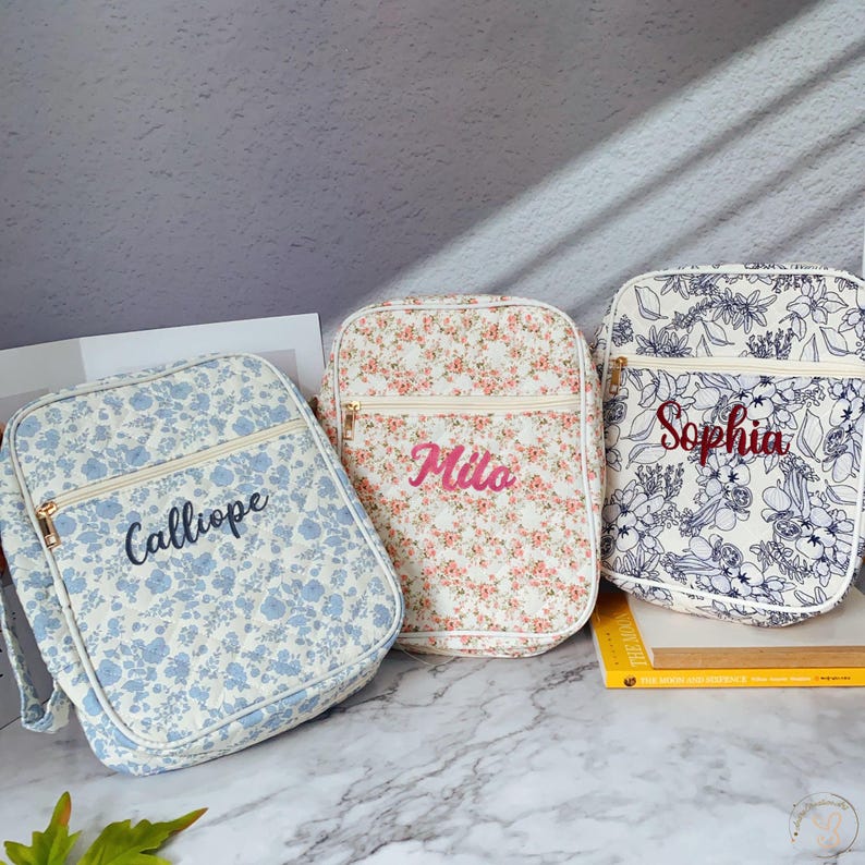 May include: Three floral pattern bags with personalized names. The bags feature a rectangular shape with a zippered front pocket and a top zipper. The bag on the left has a blue floral pattern with the name "Calliope". The middle bag has a pink floral pattern with the name "Mila". The bag on the right has a white and blue floral pattern with the name "Sophia".