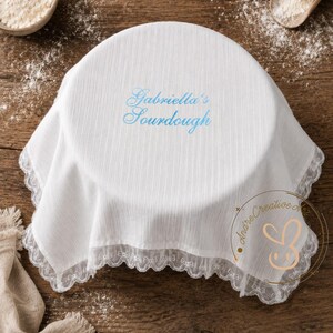 Puede incluir: Una cubierta de tela blanca circular con una textura de rayas verticales sutiles y un borde de encaje blanco delicado. El centro presenta el texto bordado "Gabriella's Sourdough" en una fuente cursiva azul claro, dise&ntilde;ada para cubrir un cuenco.