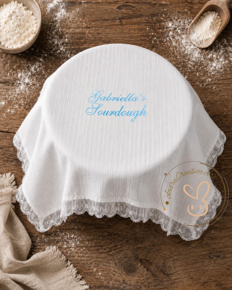 Puede incluir: Una cubierta de tela blanca circular con una textura de rayas verticales sutiles y un borde de encaje blanco delicado. El centro presenta el texto bordado "Gabriella's Sourdough" en una fuente cursiva azul claro, dise&ntilde;ada para cubrir un cuenco.