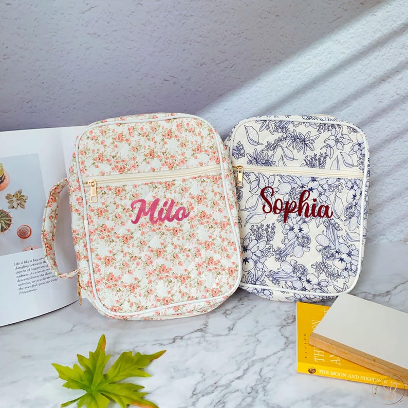 May include: Two floral print backpacks. One is white with pink flowers and the name "Milo" embroidered in pink. The other is white with blue floral designs and the name "Sophia" embroidered in red. Both have gold zippers.