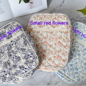 May include: Three quilted bags with floral patterns. One bag features dark blue flowers, another has small red flowers, and the third displays light blue florals. Each bag has a cream-colored zipper and a matching strap.