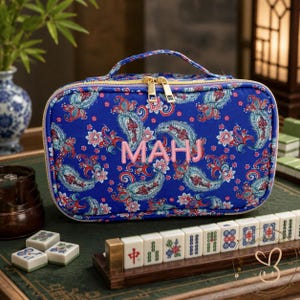 May include: A rectangular blue fabric carrying case with a top handle and gold-tone double zippers. The case features a colourful paisley pattern and the embroidered pink text MAHJ on the front. It is designed for storing game tiles.