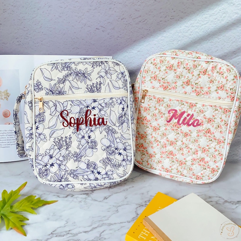 May include: Two quilted shoulder bags. One bag features a white background with a blue floral pattern and the name "Sophia" embroidered in red. The other bag has a white background with a pink floral pattern and the name "Milo" embroidered in pink. Both bags have gold zippers.