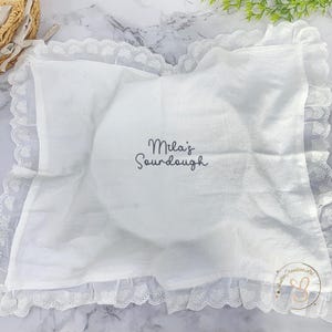 Puede incluir: Un pa&ntilde;o rectangular blanco con un borde de encaje con volantes y un patr&oacute;n de corazones. El centro est&aacute; bordado con el texto gris oscuro "Mila's Sourdough". En la esquina hay un peque&ntilde;o logotipo circular con el texto "AndreCreativeArt".
