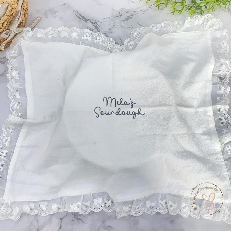 Puede incluir: Un pa&ntilde;o rectangular blanco con un borde de encaje con volantes y un patr&oacute;n de corazones. El centro est&aacute; bordado con el texto gris oscuro "Mila's Sourdough". En la esquina hay un peque&ntilde;o logotipo circular con el texto "AndreCreativeArt".
