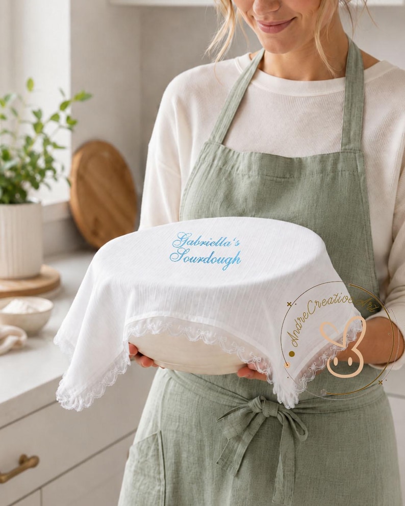 Puede incluir: Una funda de tela blanca para cuenco con un borde de encaje delicado, que presenta el texto bordado en azul Gabriella's Sourdough en la parte superior. La funda est&aacute; colocada sobre un cuenco redondo, mostrando un material texturizado y ligero.