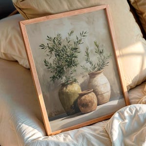 Puede incluir: Una impresión artística enmarcada que presenta una naturaleza muerta de tres jarrones de cerámica con ramas de olivo. Los jarrones son en tonos verdes, beige y marrón. La obra de arte está sobre un fondo neutro y el marco es marrón claro.