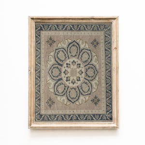 May include: A framed, rectangular rug with a floral design. The rug features a central flower motif in shades of blue, beige, and brown, surrounded by a dark blue border. The frame is made of light-colored wood, adding a vintage aesthetic.