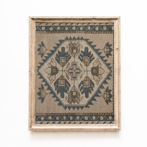 May include: A framed textile art piece with a geometric design. The artwork features a central diamond shape with a cross and floral motifs in shades of blue and brown on a beige background. The frame is made of light-colored wood.