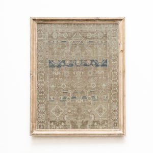 Op de afbeelding: Een ingelijste print van een vintage tapijt met een verweerde uitstraling. Het tapijt heeft een gedetailleerd patroon in beige, blauwe en bruine tinten. De lijst is gemaakt van lichtgekleurd hout, wat het kunstwerk een rustieke uitstraling geeft.