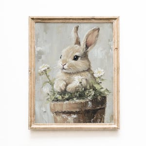 May include: A framed painting of a tan bunny in a brown flower pot with white flowers. The bunny has long ears and dark eyes. The painting has a light gray background and a light brown wooden frame.