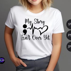 Puede incluir: Camiseta blanca con el texto negro "My Story Isn't Over Yet". El dise&ntilde;o incluye un punto y coma, una l&iacute;nea de latidos y un coraz&oacute;n. La camiseta es de cuello redondo.