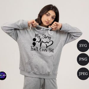 Puede incluir: Una sudadera con capucha gris claro con el texto negro "My Story Isn't Over Yet". El dise&ntilde;o incluye un punto y coma, una l&iacute;nea de latidos y un coraz&oacute;n. La sudadera tiene un bolsillo delantero y una capucha con cord&oacute;n.