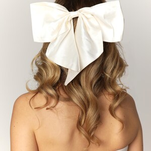 May include: A large, white satin bow hair accessory. The bow is tied in a large, loose knot and has long, flowing tails.