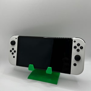 May include: A green plastic stand for a Nintendo Switch console. The stand is designed to hold the console upright, allowing for easy viewing of the screen.