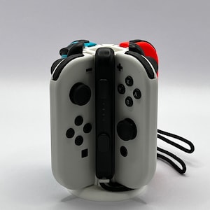 May include: A white charging dock with a black Nintendo Switch console and attached Joy-Con controllers. The controllers are white, red, and blue. The dock has a circular base and is set against a white background.