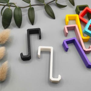Peut inclure: Un ensemble de 10 crochets en plastique de différentes couleurs, dont noir, blanc, violet, rose, jaune, bleu et vert. Les crochets sont en forme de 'J' inversé et sont conçus pour accrocher des objets à un mur ou une porte.