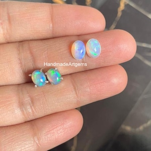 May include: A close-up of a hand holding two oval-shaped opals and a pair of opal stud earrings. The opals display a mix of blue, yellow, and green hues. The earrings have a silver setting. The text "HandmadeArtgems" is visible.