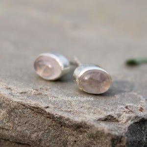 Op de afbeelding: Een paar oorknopjes met ovale, lichtroze rozenkwarts edelstenen gezet in gladde, zilverkleurige metalen zettingen. De tekst HandmadeArtgems is zichtbaar in het midden onderaan de afbeelding, gepositioneerd tussen de twee oorbellen.