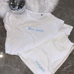 Może przedstawiać: Biały bawełniany zestaw piżamy z niebieskim haftem. Góra ma napis "Mrs. Jones", a spodenki "14.08.2025".