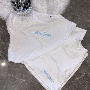 Puede incluir: Conjunto de pijama de algod&oacute;n blanco con bordado azul. La parte superior dice "Mrs. Jones" y los pantalones cortos dicen "14.08.2025".