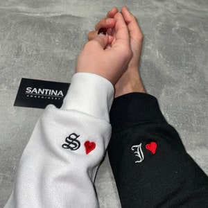 May include: Two white and black sweatshirts with embroidered red hearts and the letters "S" and "J" on the sleeves. The sweatshirts are held together by two hands.