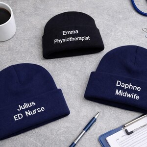 Könnte beinhalten: Drei Mützen, zwei marineblaue und eine schwarze, werden auf einer grauen Oberfläche gezeigt. Die schwarze Mütze hat die weiße Aufschrift "Emma Physiotherapeut". Die marineblauen Mützen tragen die Aufschrift "Julius ED Nurse" und "Daphne Midwife". Eine Kaffeetasse, ein Stethoskop und ein Stift sind ebenfalls zu sehen.