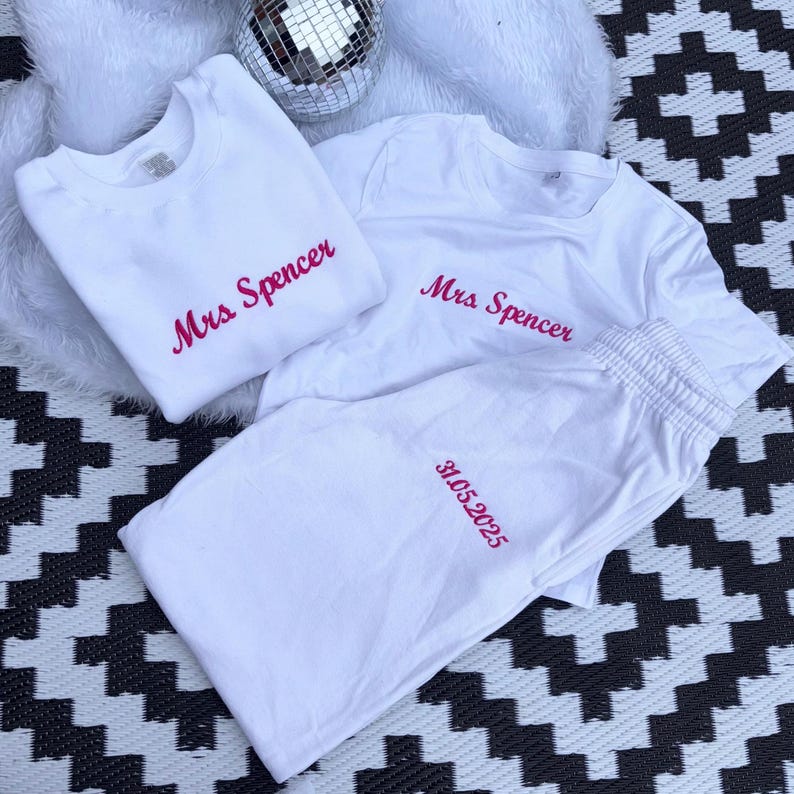 May include: White sweatshirt, t-shirt, and sweatpants set. The text "Mrs Spencer" is embroidered in pink on the sweatshirt and t-shirt. The sweatpants have the date "31.05.2025" embroidered in pink. A disco ball is in the background.